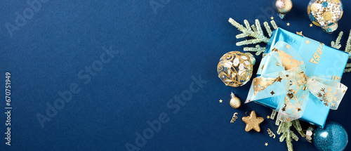 Christmas gift box with fir branches, gold Xmas ball ornaments, decorations, confetti on dark blue background. Christmas banner design