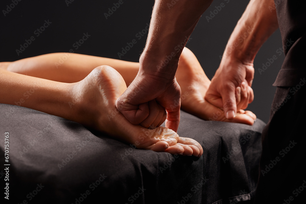 handsome male masseur doing a massage on a girl's leg on a black background, concept of therapeutic relaxing massage - obrazy, fototapety, plakaty 