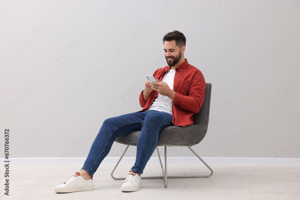 Handsome man using smartphone while sitting in armchair near light grey wall indoors Handsome man using smartphone while sitting in armchair near light grey wall indoors