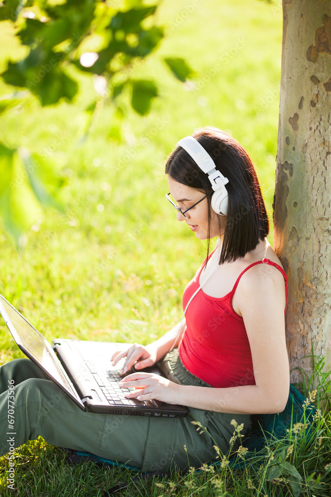 girl with dark brown hair wearing white headphones using laptop in a park under a tree girl with dark brown hair wearing white headphones using laptop in a park under a tree
