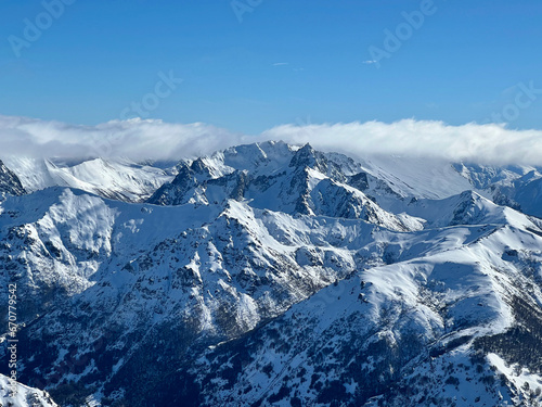 A high-angle view of a snowy mountain range in Cerro Catedral. The sky is blue and the clouds are low. The mountains are jagged and textured. © Gerardo