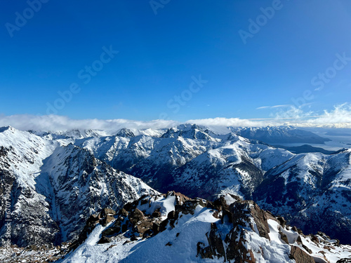 A high-angle view of snowy mountains and a blue lake in Cerro Catedral. The sky is blue with some clouds. The mountains are jagged and rocky.