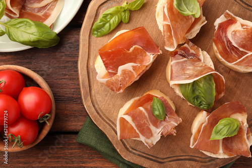 Board of tasty sandwiches with cured ham and basil leaves near tomatoes on wooden table, flat lay