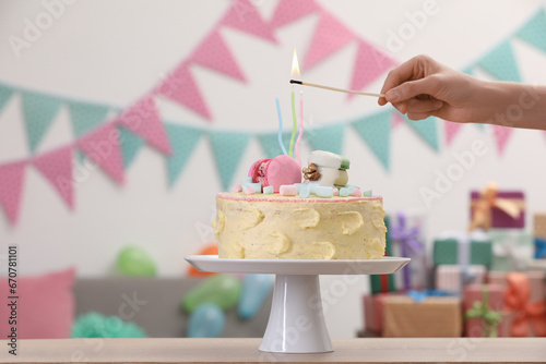 Woman lighting candle on delicious cake decorated with macarons and marshmallows in festive room, closeup. Space for text