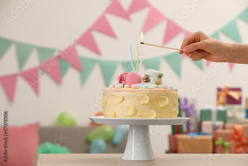 Woman lighting candle on delicious cake decorated with macarons and marshmallows in festive room, closeup. Space for text
