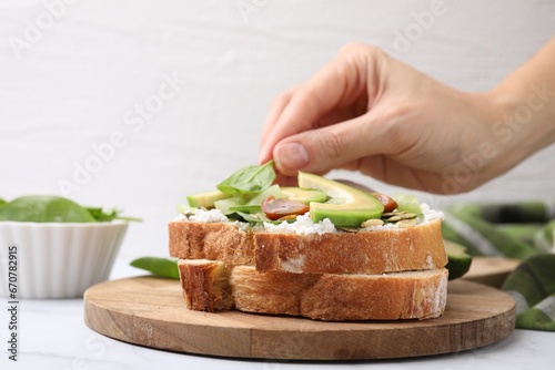 Woman putting spinach leaf on tasty vegan sandwich with avocado and tomato at white marble table, closeup