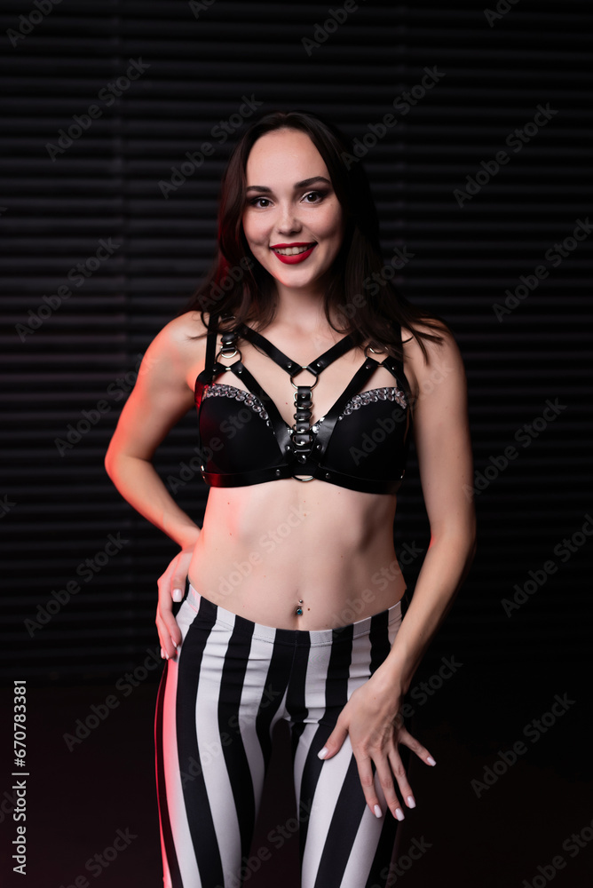 Portrait of a beautiful girl in a black bra, leather belts and leggings in black and white lines posing against the background of a wooden wall. Back view. Party concept, night club