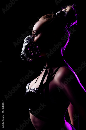 Portrait of a beautiful girl in black underwear and leather harness. On the face is a respirator decorated with rhinestones. Contour purple light. Party concept, night club © Granmedia