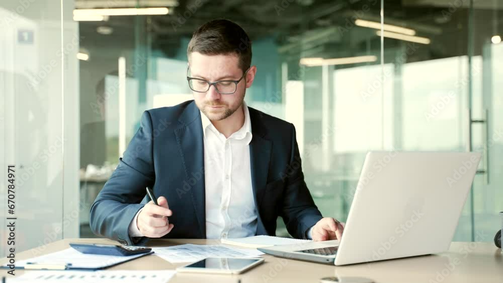 Busy financier in formal suit making financial calculations using calculator, typing on a laptop taking notes sitting at workplace. Confident businessman working fills out documents in business office Busy financier in formal suit making financial calculations using calculator, typing on a laptop taking notes sitting at workplace. Confident businessman working fills out documents in business office