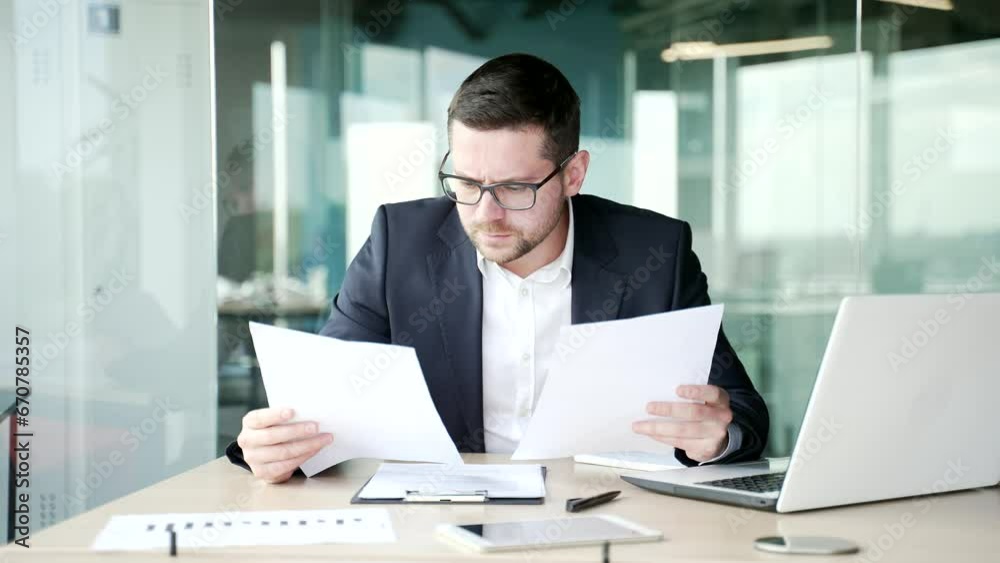 Disappointed businessman in formal suit having difficulty with paper work sitting at workplace in business office. Confused disgruntled financier holding documents, doesn't understand the problem