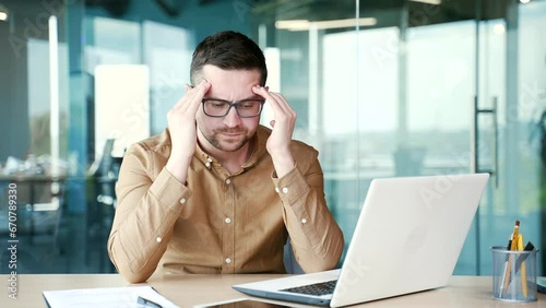 Wallpaper Mural Young businessman suffering from a headache while working on a laptop while sitting at workplace in office. Sad worker massaging his temples with his hands. He is exhausted and has high blood pressure Torontodigital.ca