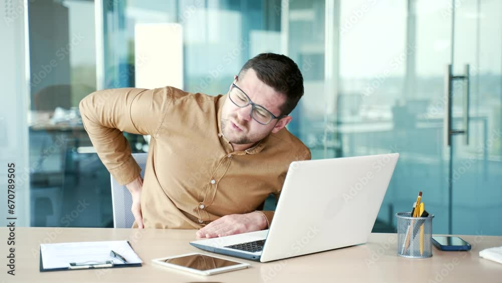 Tired young businessman suffering from back pain while working on laptop sitting at desk at workplace in business office. Worker massages painful lower back muscles. He is exhausted and overworked Tired young businessman suffering from back pain while working on laptop sitting at desk at workplace in business office. Worker massages painful lower back muscles. He is exhausted and overworked