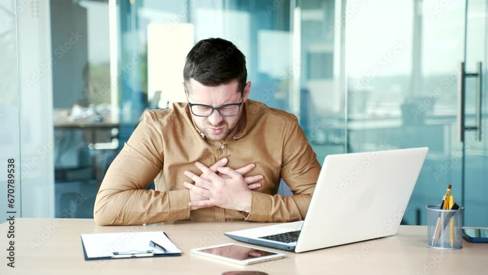 Young businessman has a heart attack while working on a laptop while sitting at a workplace in a business office. A sick worker suffers from tension in the chest and holds the sore spot with his hands Young businessman has a heart attack while working on a laptop while sitting at a workplace in a business office. A sick worker suffers from tension in the chest and holds the sore spot with his hands