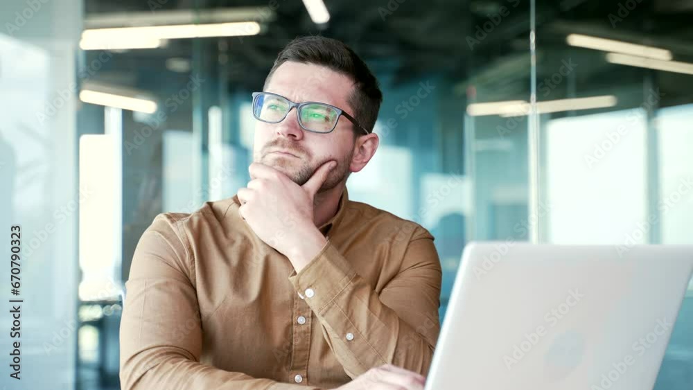 Serious thoughtful businessman in a shirt thinks while working on a laptop sitting at workplace in a business office. A confident entrepreneur in glasses is busy with a startup project on the computer Serious thoughtful businessman in a shirt thinks while working on a laptop sitting at workplace in a business office. A confident entrepreneur in glasses is busy with a startup project on the computer