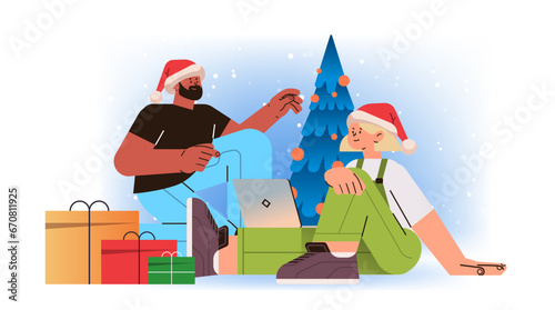mix race man woman in santa hats near decorated christmas tree using laptop social media communication new year holidays celebration concept