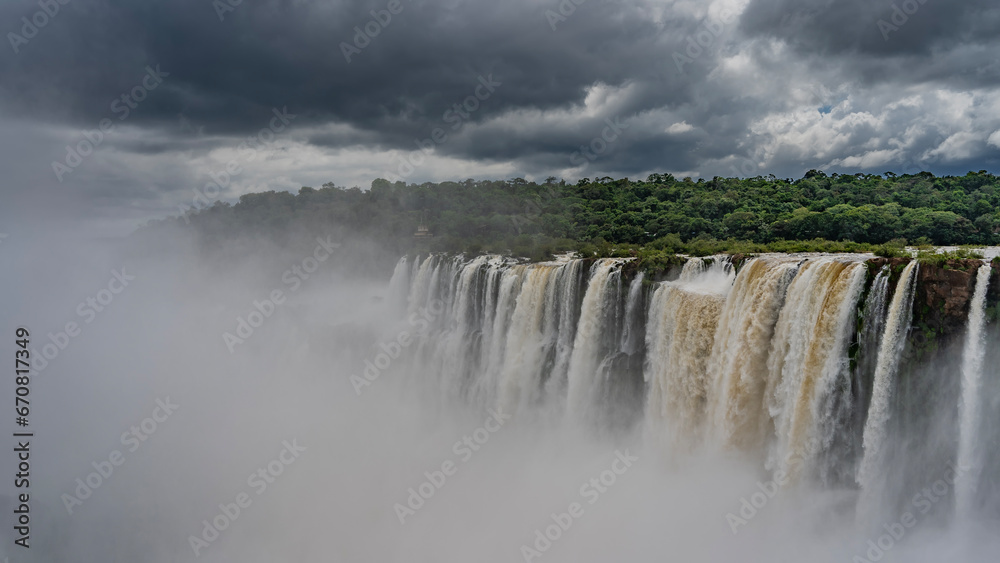 Beautiful waterfall landscape. Streams of water cascade into the abyss from the ledge. Lush green vegetation on the riverbank. Splashes, fog in the foreground. Iguazu Falls. Argentina. Beautiful waterfall landscape. Streams of water cascade into the abyss from the ledge. Lush green vegetation on the riverbank. Splashes, fog in the foreground. Iguazu Falls. Argentina.