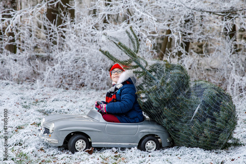 Happy little smiling girl driving toy car with Christmas tree. Funny preschool child in winter clothes bringing hewed xmas tree from snowy forest. Family, tradition, holiday. photo