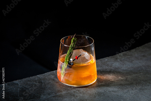 orange alcoholic cocktail with a sprig of pine needles and round ice, side view