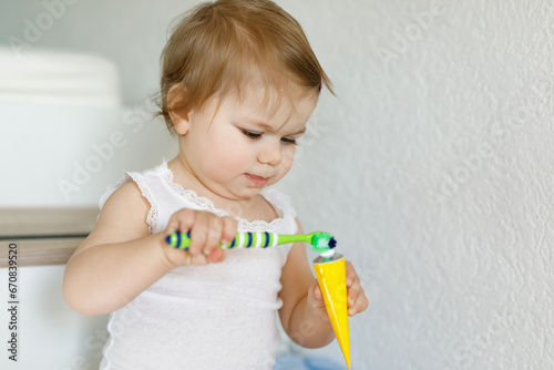 Little baby girl holding toothbrush and brushing first teeth. Toddler learning to clean milk tooth. Prevention, hygiene and healthcare concept. Happy child in bathroom