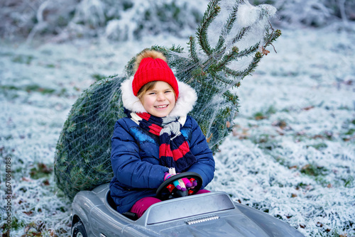 Happy little smiling girl driving toy car with Christmas tree. Funny preschool child in winter clothes bringing hewed xmas tree from snowy forest. Family, tradition, holiday. photo