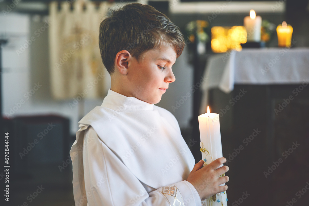 Little kid boy receiving his first holy communion. Happy child holding Christening candle. Tradition in catholic curch. Kid in a white traditional gown in a church near altar.
