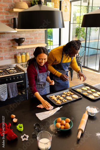 Happy diverse couple in aprons preparing christmas cookies and cleaning in kitchen, copy space
