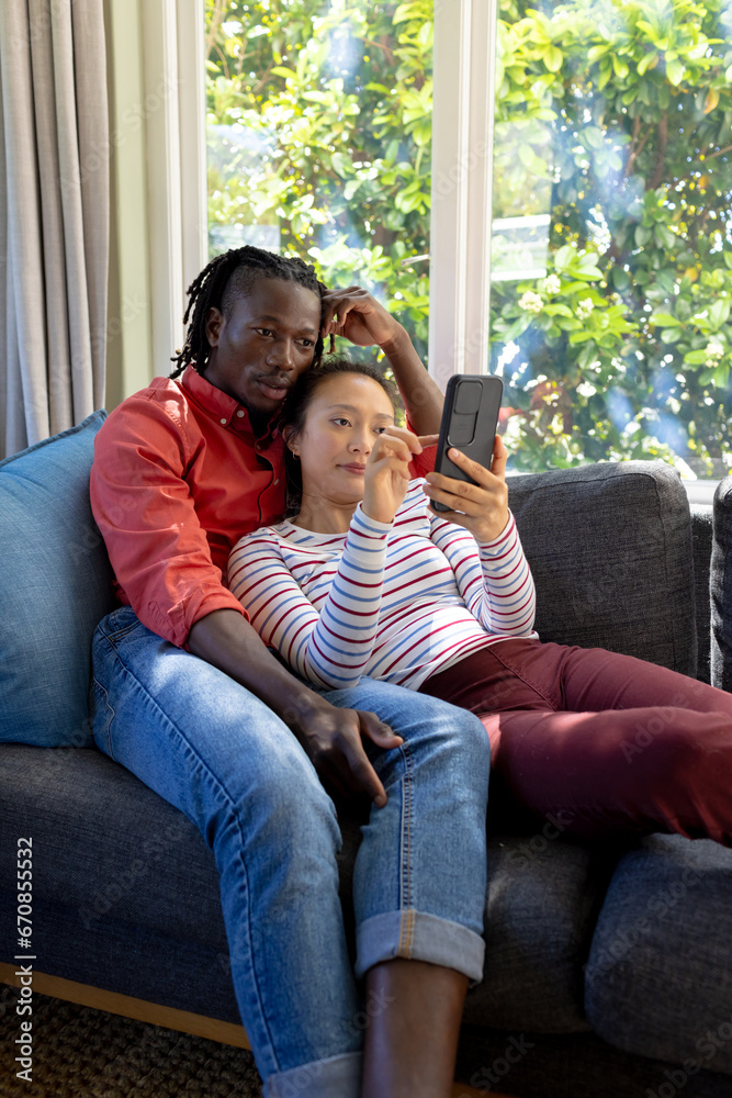 Obraz premium Focused diverse couple using smartphone relaxing on couch in sunny living room, copy space