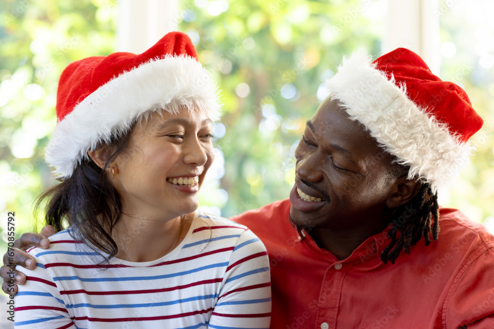 Happy diverse couple in christmas hats embracing and smiling at each other in sunny living room