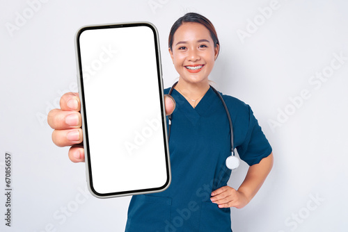 Smiling young Asian woman nurse working wearing a blue uniform showing smartphone with blank screen isolated on white background. Healthcare medicine concept
