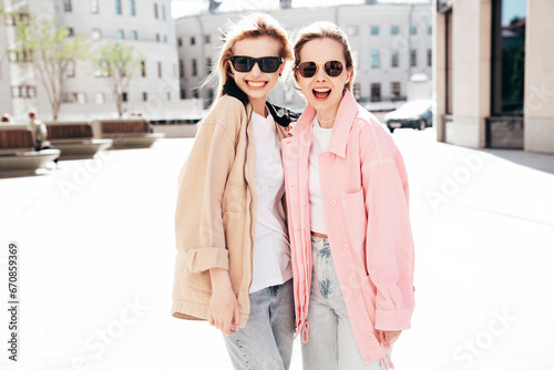 Two young beautiful smiling hipster female in trendy summer clothes. Carefree women posing in the street. Positive models having fun outdoors at sunny day. Cheerful and happy. In sunglasses