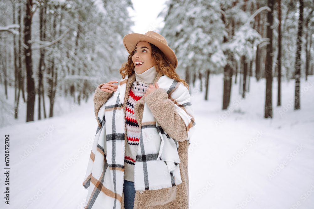 Fototapeta premium Happy woman among snowy trees in winter forest, enjoying the first snow. A young woman in a hat and scarf is having fun in the winter forest. Lifestyle.