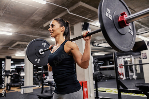 A happy strong woman is lifting barbell in a gym and doing exercises for shoulders.