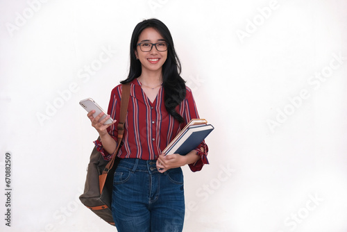 Female student standing alone holding her smartphone while carrying some books and school bag photo