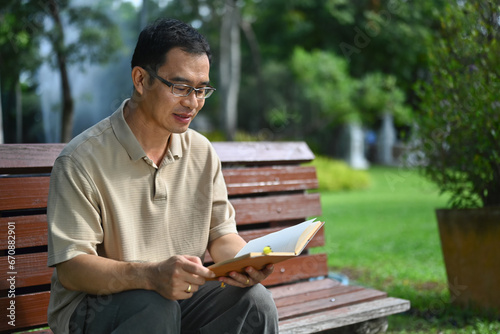 Pleased middle age man reading favorite novel while sitting on bench at public park