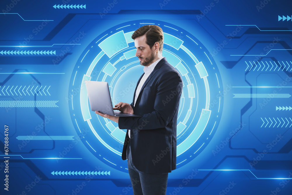 Attractive young businessman using laptop on creative blue hi-tech background. Technology, future and innovation concept.