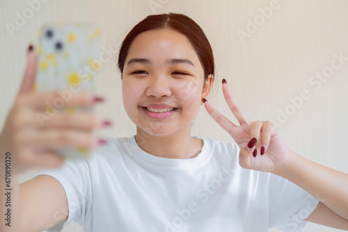 Image of beautiful Asian girl laughing and showing peace sign while taking selfie photo on cellphone.