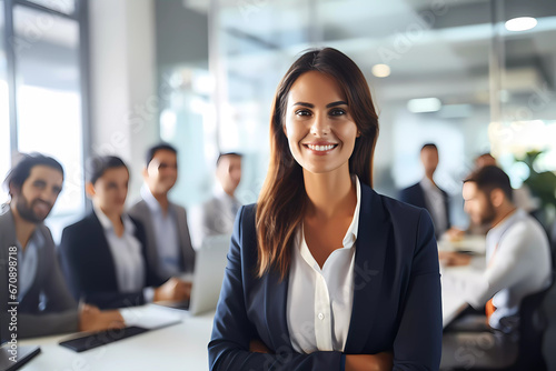 Portrait of beautiful brunette hair businesswoman in a busy modern workplace, standing in front of the team, smiling at camera. Office employee posing with coworkers in background. 