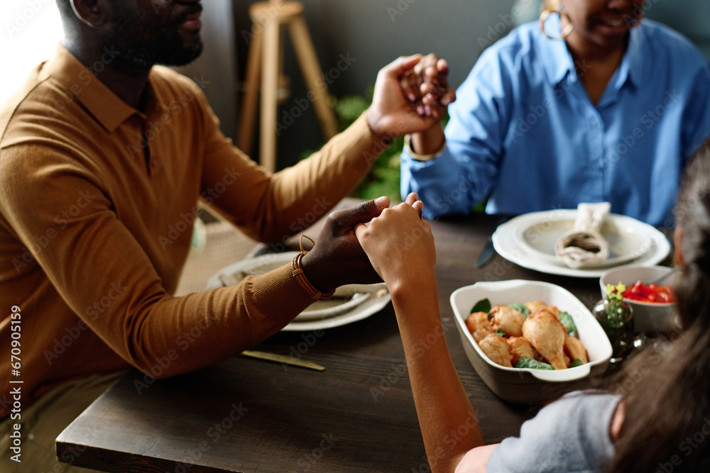 Focus on hands of family members giving thanks to Lord on holiday while sitting by table served with homemade food and enjoying festive dinner Focus on hands of family members giving thanks to Lord on holiday while sitting by table served with homemade food and enjoying festive dinner