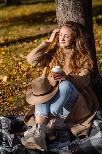 Beautiful blonde woman in a coat and hat sits on a plaid under a tree in an autumn park on a sunny day
