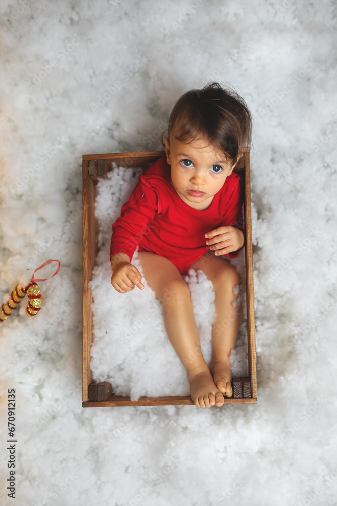 Fototapeta premium Young kid in red lying in wooden box surrounded with snow. Christmas and New Year scenography studio photo. 
