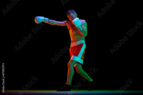 One man, professional boxer in gloves training against over black mode background in mixed neon filter, light. © Lustre