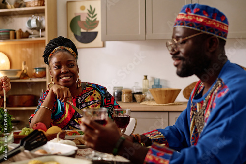 Happy African American woman in ethnic apparel looking at her husband with smile while sitting in front of him by served festive table at home photo