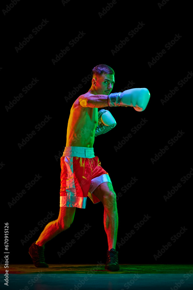 Talented shirtless boxer, mixed martial art fighter exercising before fight against black mode background in mixed neon filter, light.