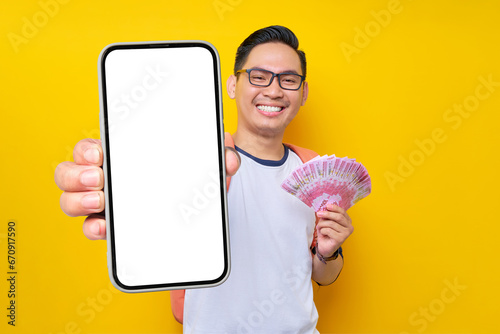 Smiling young Asian student man in casual clothes and glasses backpack holding smartphone blank screen and cash money isolated on yellow background. Education in High School University College concept photo
