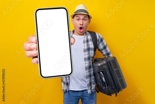 Surprised young traveler tourist Asian man in casual clothes hat with backpack carrying suitcase in hand and mobile phone isolated on yellow background. Air flight journey concept photo
