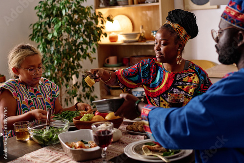 Young African American woman passing baked corn to her daughter by festive table served with homemade food for kwanzaa dinner