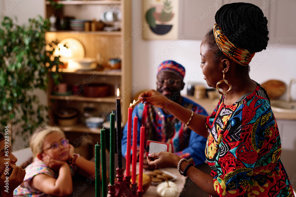 Side view of young African American woman in national dress and accessories burning candles symbolizing seven kwanzaa principles - obrazy, fototapety, plakaty 