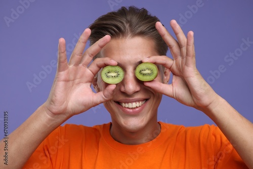 Smiling man covering eyes with halves of kiwi on violet background, closeup