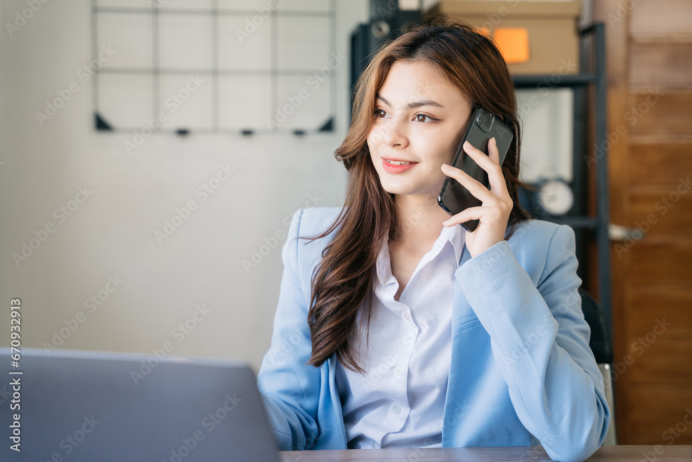 businesswoman talking mobile phone and working on laptop and papers on table in her office.