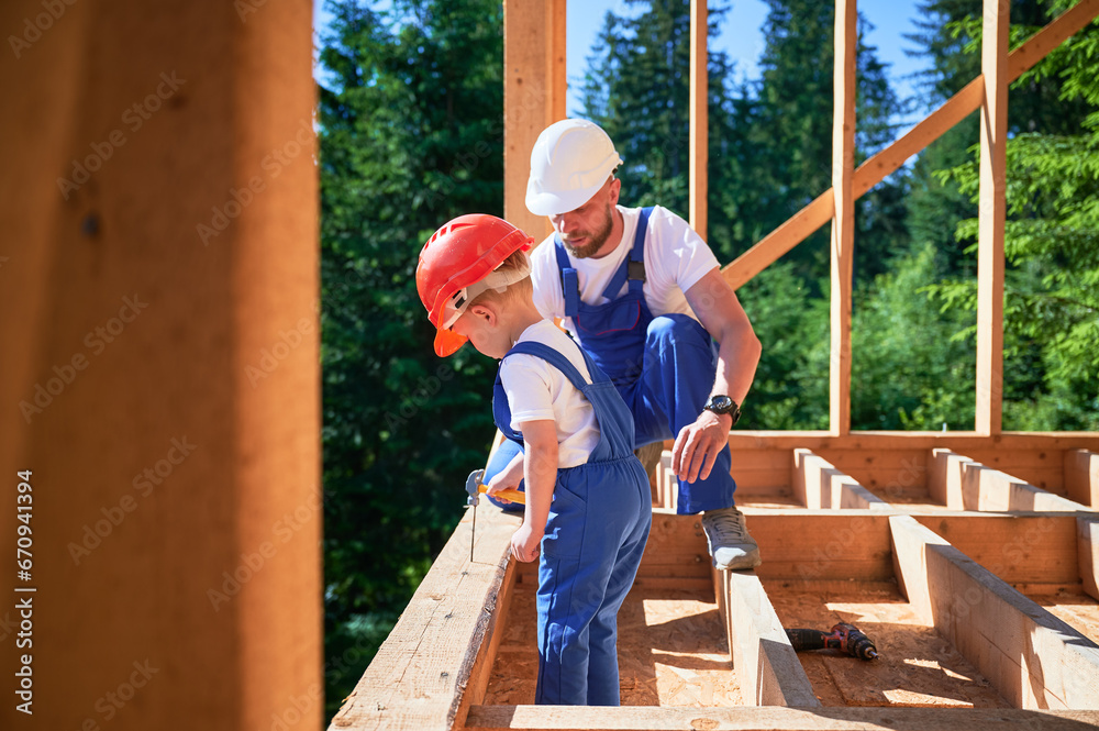 Father with toddler son constructing wooden frame house. Father construction worker imparting the skill of driving nails with hammer to his son on sunny day. Carpentry and family concept. Father with toddler son constructing wooden frame house. Father construction worker imparting the skill of driving nails with hammer to his son on sunny day. Carpentry and family concept.